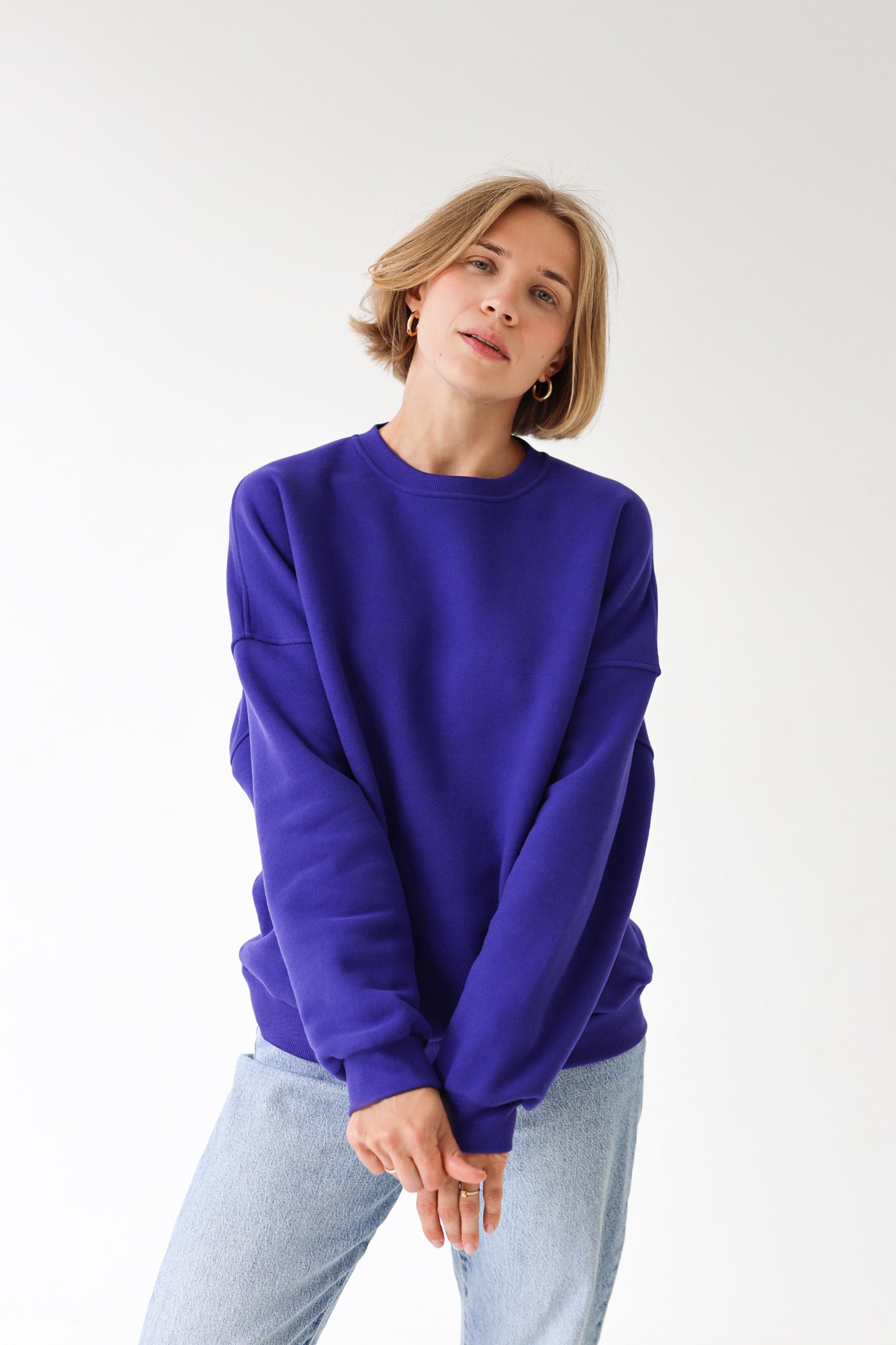 Portret kobiety w fioletowej bluzie i jeansach na białym tle. Krótkie blond włosy, złote kolczyki koła. Naturalne światło, minimalistyczny styl.