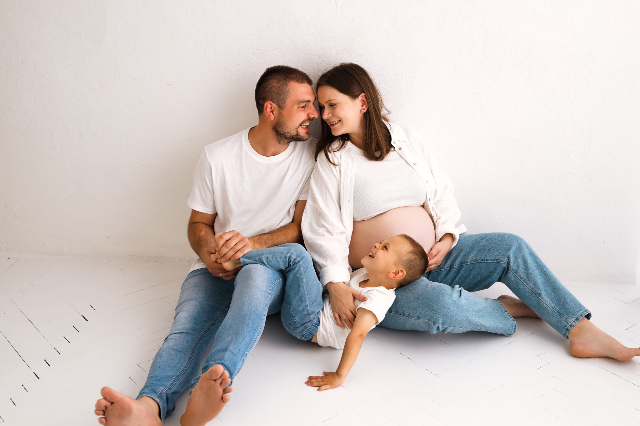 Uśmiechnięta, przyszła mama z partnerem i synkiem w jasnym studiu. Naturalne światło podkreśla więź rodziny, ubranych w jeans i białe koszulki.