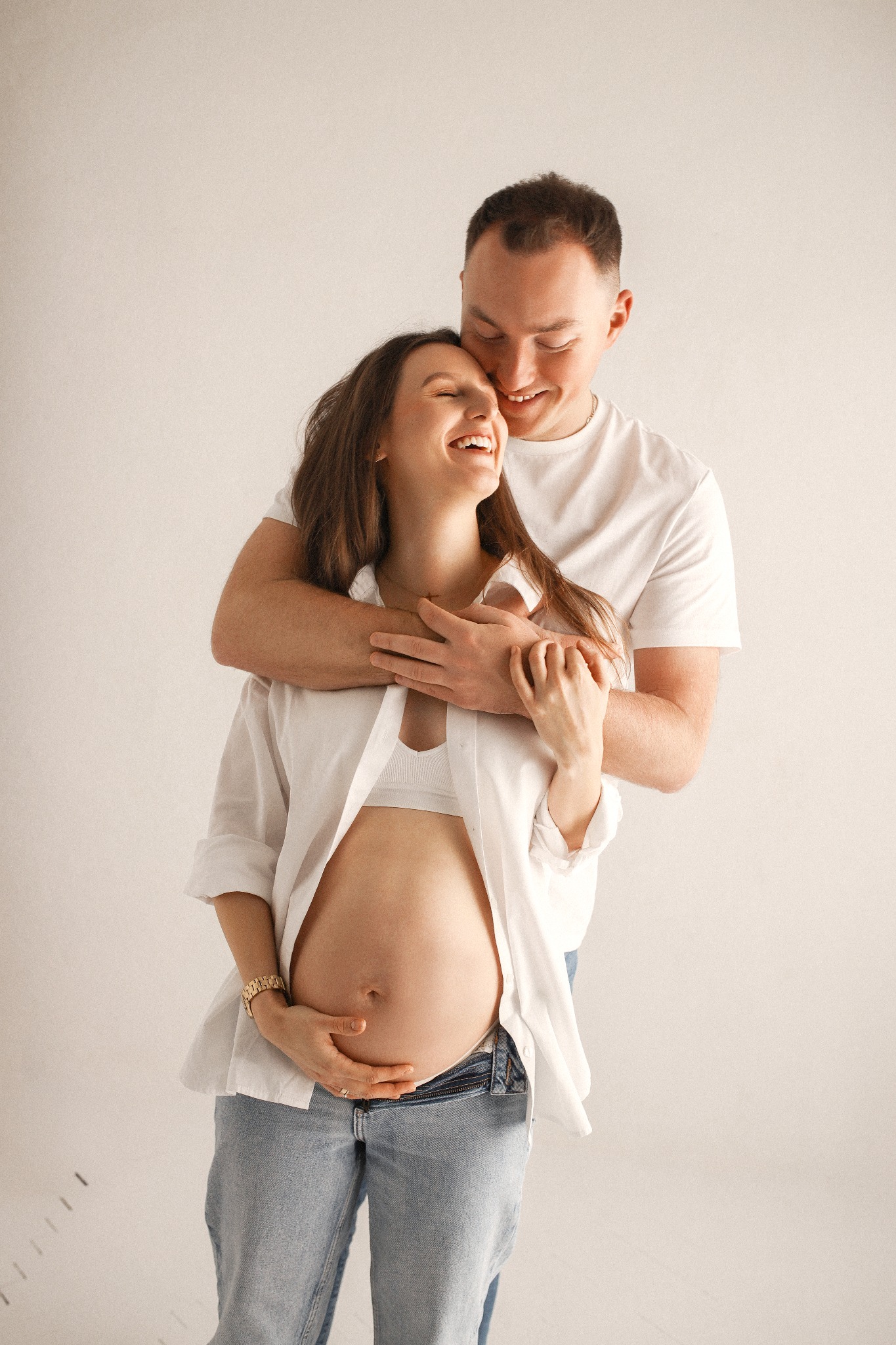 Uśmiechnięta, brzemienna kobieta w jasnej koszuli i jeansach, obejmowana przez mężczyznę w białym T-shircie na neutralnym tle. Delikatne światło podkreśla radość oczekiwania.