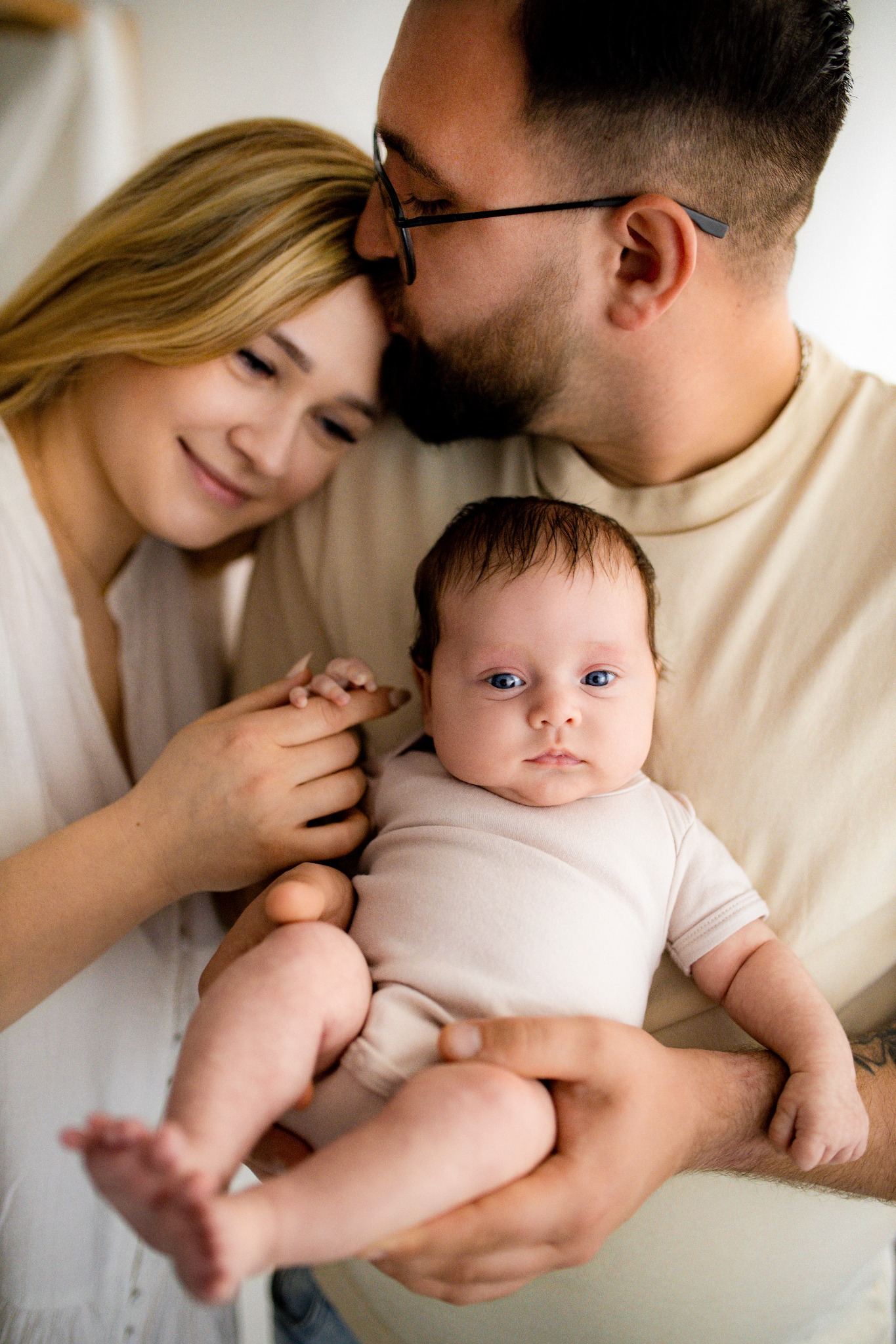 Rodzinne ujęcie: ojciec w okularach całuje matkę w czoło, trzymając na rękach niemowlę z intensywnym spojrzeniem, w jasnym wnętrzu.