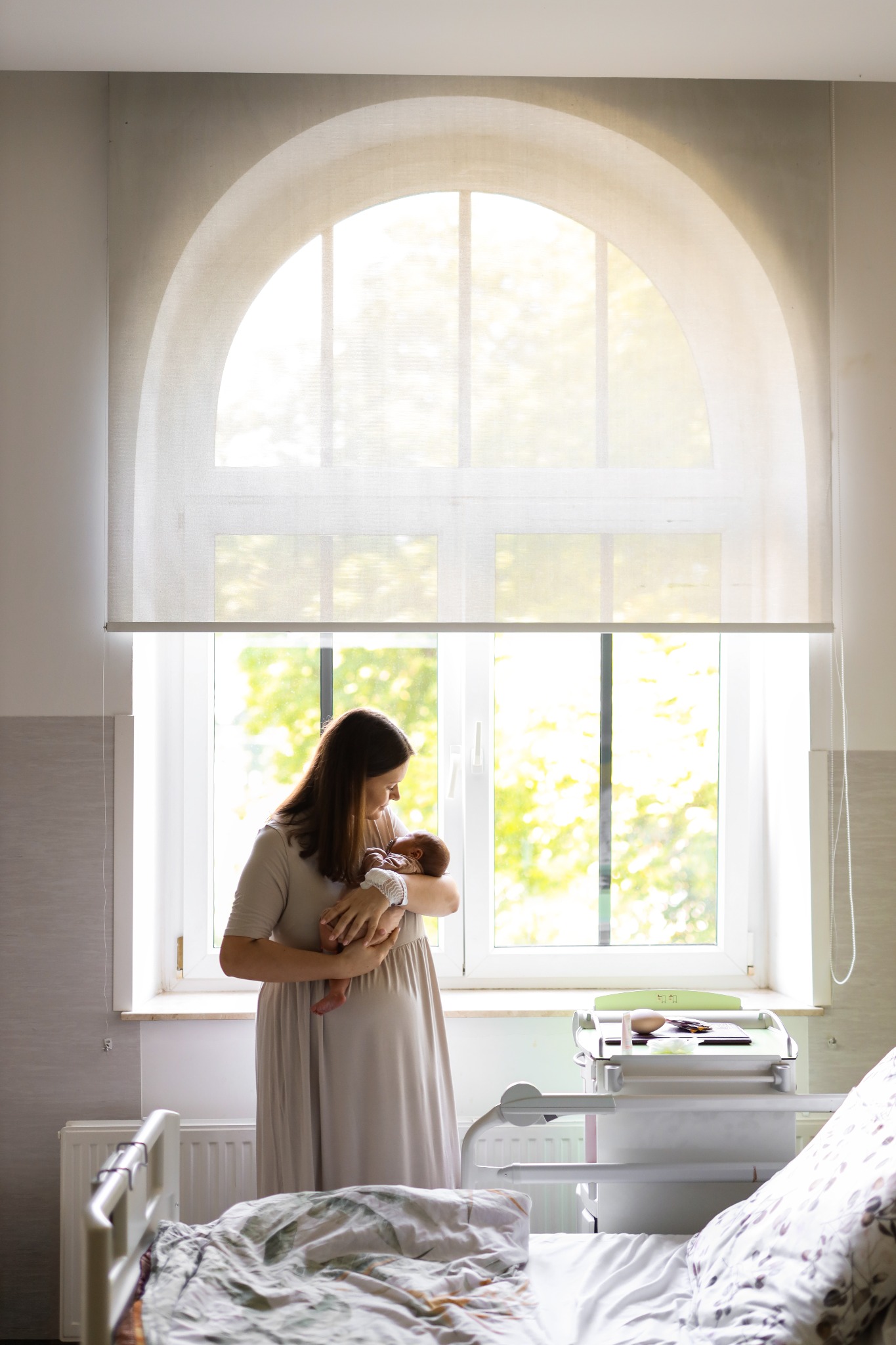 Mama trzyma noworodka w szpitalnej sali z dużym oknem łukowym. Delikatne światło podkreśla intymność i spokój. Widoczny wózek szpitalny i łóżko.