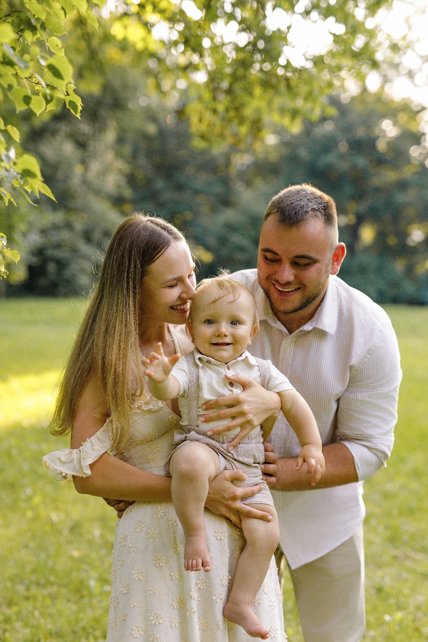 Uśmiechnięta rodzina w parku: mama w kwiecistej sukience trzyma synka, tata obejmuje ich. Delikatne światło podkreśla radosną atmosferę sesji rodzinnej.