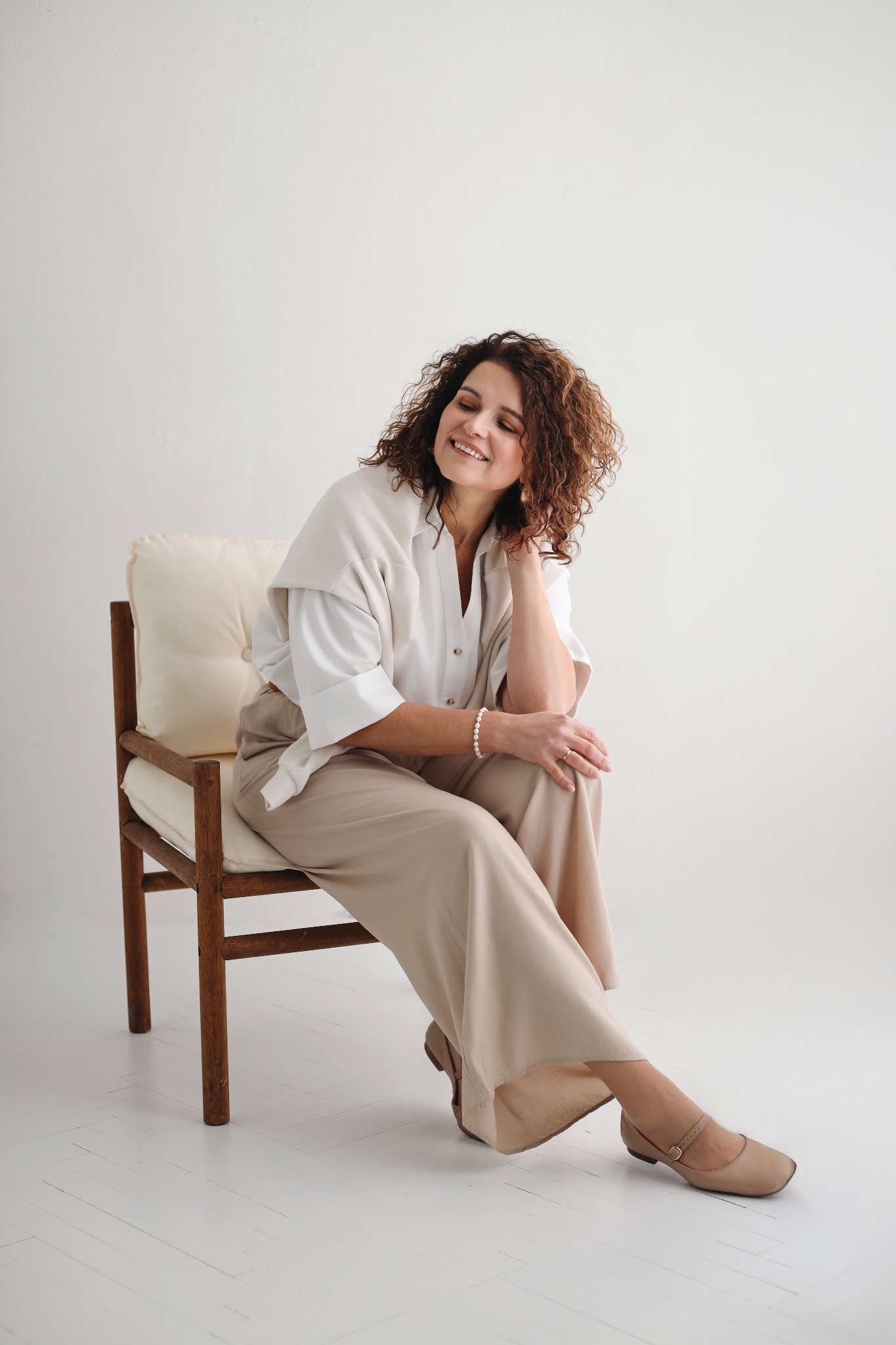 Kobieta z kręconymi włosami siedzi na krześle w jasnym wnętrzu, ubrana w beżowe spodnie i białą koszulę. Uśmiech na twarzy, delikatna biżuteria.