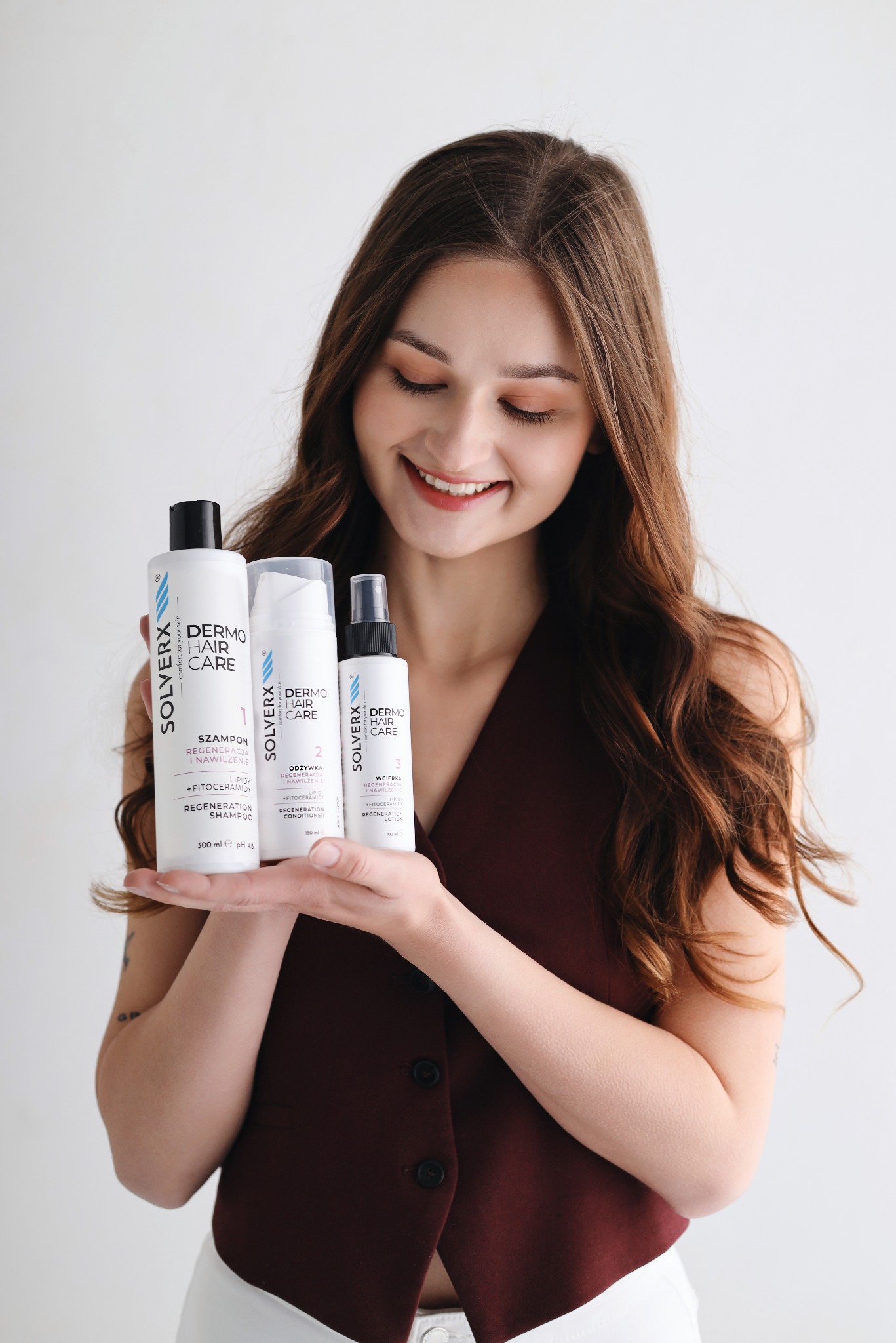 Uśmiechnięta brunetka prezentuje zestaw kosmetyków Solverx Dermo Hair Care: szampon, odżywkę i wcierkę. Jasne tło, profesjonalna fotografia produktowa.