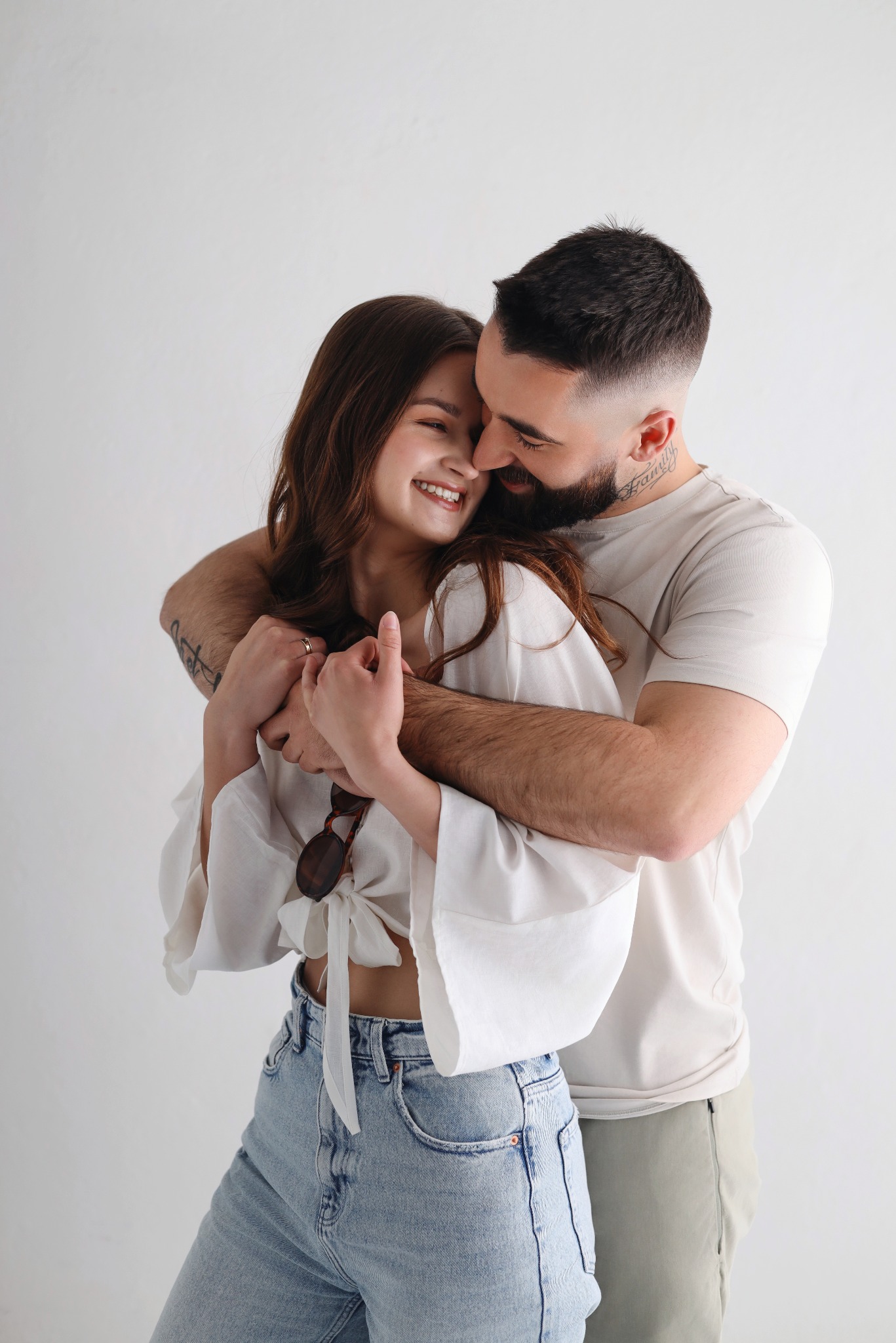 Uśmiechnięta para w objęciach, ona w białej bluzce i jeansach, on w beżowej koszulce, na jasnym tle. Widoczne tatuaże, okulary i pierścionek.