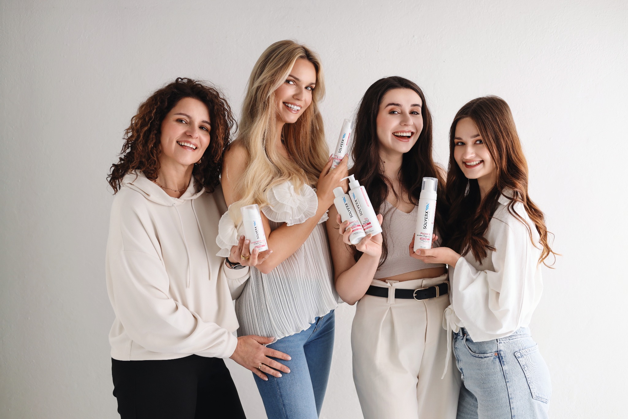 Cztery uśmiechnięte kobiety prezentują kosmetyki Solverx na białym tle. Różne typy urody, jasne ubrania, naturalne światło. Zdjęcie reklamowe.