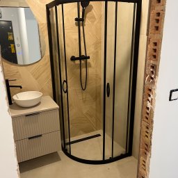 Filip Szumny Nieruchomości - Nowoczesna kabina prysznicowa z czarną armaturą i umywalką na szafce w łazience z beżowymi płytkami. Widoczne surowe wykończenie ściany.