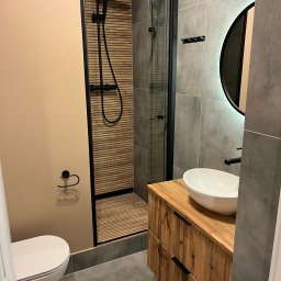 Filip Szumny Nieruchomości - Nowoczesna łazienka z kabiną prysznicową typu walk-in, drewnianą szafką pod umywalką i okrągłym lustrem z podświetleniem. Uwagę zwraca wykończenie ściany prysznica drewnianymi lamelami.