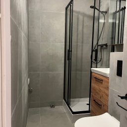 Filip Szumny Nieruchomości - Nowoczesna łazienka w szarościach z kabiną prysznicową w czarnej ramie, drewnianą szafką pod umywalką i wiszącą toaletą. Minimalistyczny design, płytki wielkoformatowe.