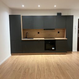 Filip Szumny Nieruchomości - Nowoczesna, szara zabudowa kuchenna z drewnianym akcentem i czarną armaturą, oświetlona punktowo, na tle drewnianej podłogi. Minimalistyczny design.