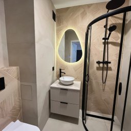 Filip Szumny Nieruchomości - Nowoczesna łazienka po remoncie: kabina prysznicowa z czarną armaturą, umywalka na szafce z szufladami i podświetlane lustro o nieregularnym kształcie, beżowe płytki.