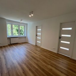 Filip Szumny Nieruchomości - Jasne wnętrze po remoncie: drewnopodobna podłoga, białe ściany, okno z widokiem na zieleń i dwoje białych drzwi z przeszkleniami. Lampy sufitowe.