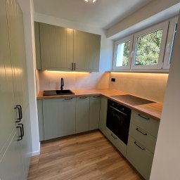 Filip Szumny Nieruchomości - Jasna kuchnia w odcieniach zieleni z drewnianym blatem, czarną armaturą i oświetleniem LED pod szafkami. Widok na okno z zielenią na zewnątrz.