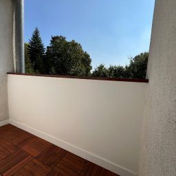 Filip Szumny Nieruchomości - Wykończony balkon z drewnianą podłogą i świeżo pomalowanymi ścianami. Widok na zieleń za balustradą. Słoneczny dzień.