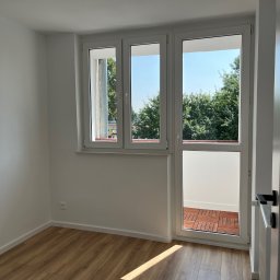 Filip Szumny Nieruchomości - Jasne wnętrze pokoju z drewnianą podłogą i białymi ścianami. Trzyczęściowe okno balkonowe z widokiem na zieleń. Nowoczesne drzwi z czarną klamką po prawej.