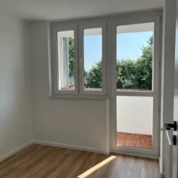 Jasne wnętrze pokoju z drewnianą podłogą i białymi ścianami. Trzyczęściowe okno balkonowe z widokiem na zieleń. Nowoczesne drzwi z czarną klamką po prawej.