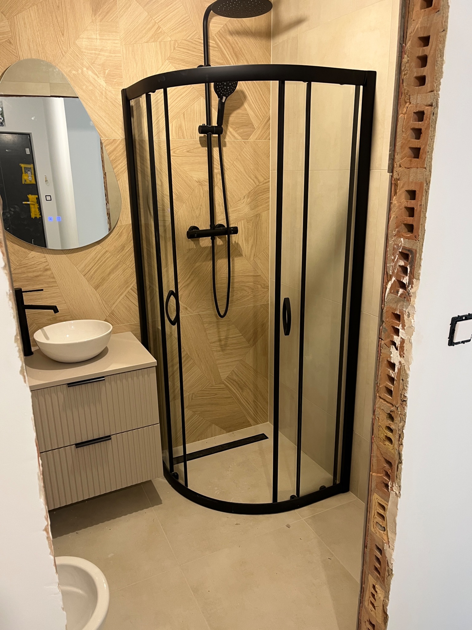 Nowoczesna kabina prysznicowa z czarną armaturą i umywalką na szafce w łazience z beżowymi płytkami. Widoczne surowe wykończenie ściany.
