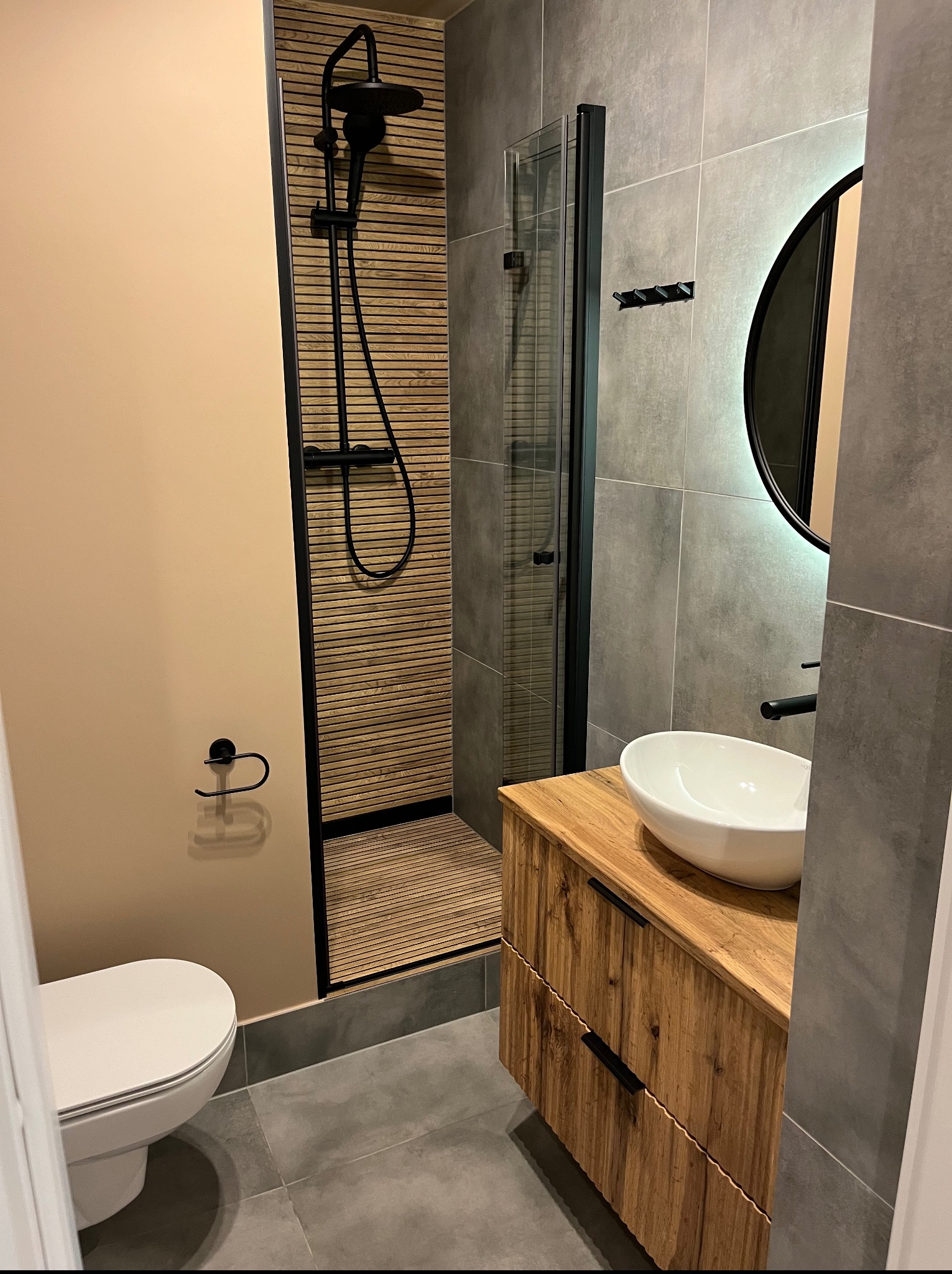 Nowoczesna łazienka z kabiną prysznicową typu walk-in, drewnianą szafką pod umywalką i okrągłym lustrem z podświetleniem. Uwagę zwraca wykończenie ściany prysznica drewnianymi lamelami.