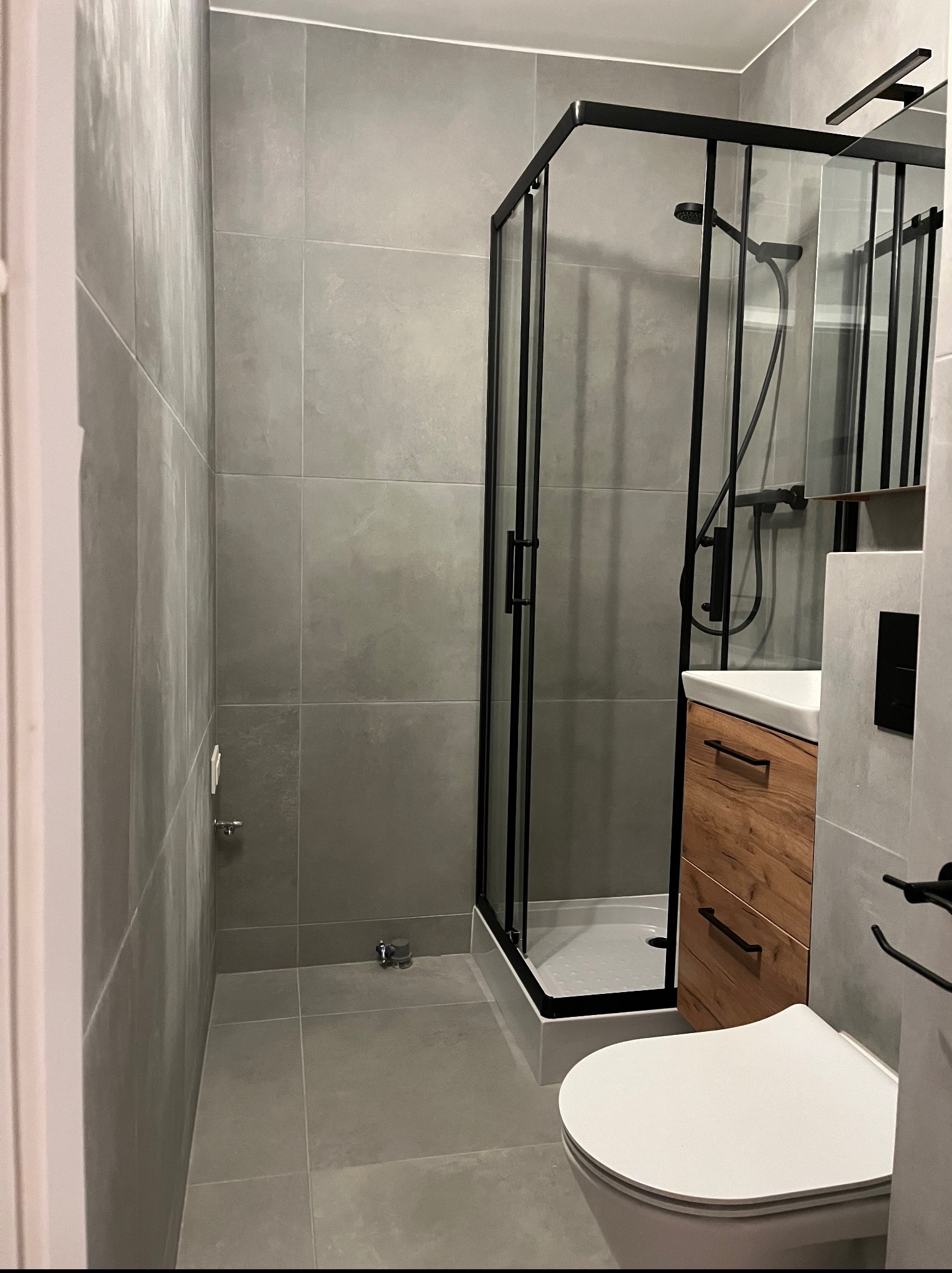 Nowoczesna łazienka w szarościach z kabiną prysznicową w czarnej ramie, drewnianą szafką pod umywalką i wiszącą toaletą. Minimalistyczny design, płytki wielkoformatowe.