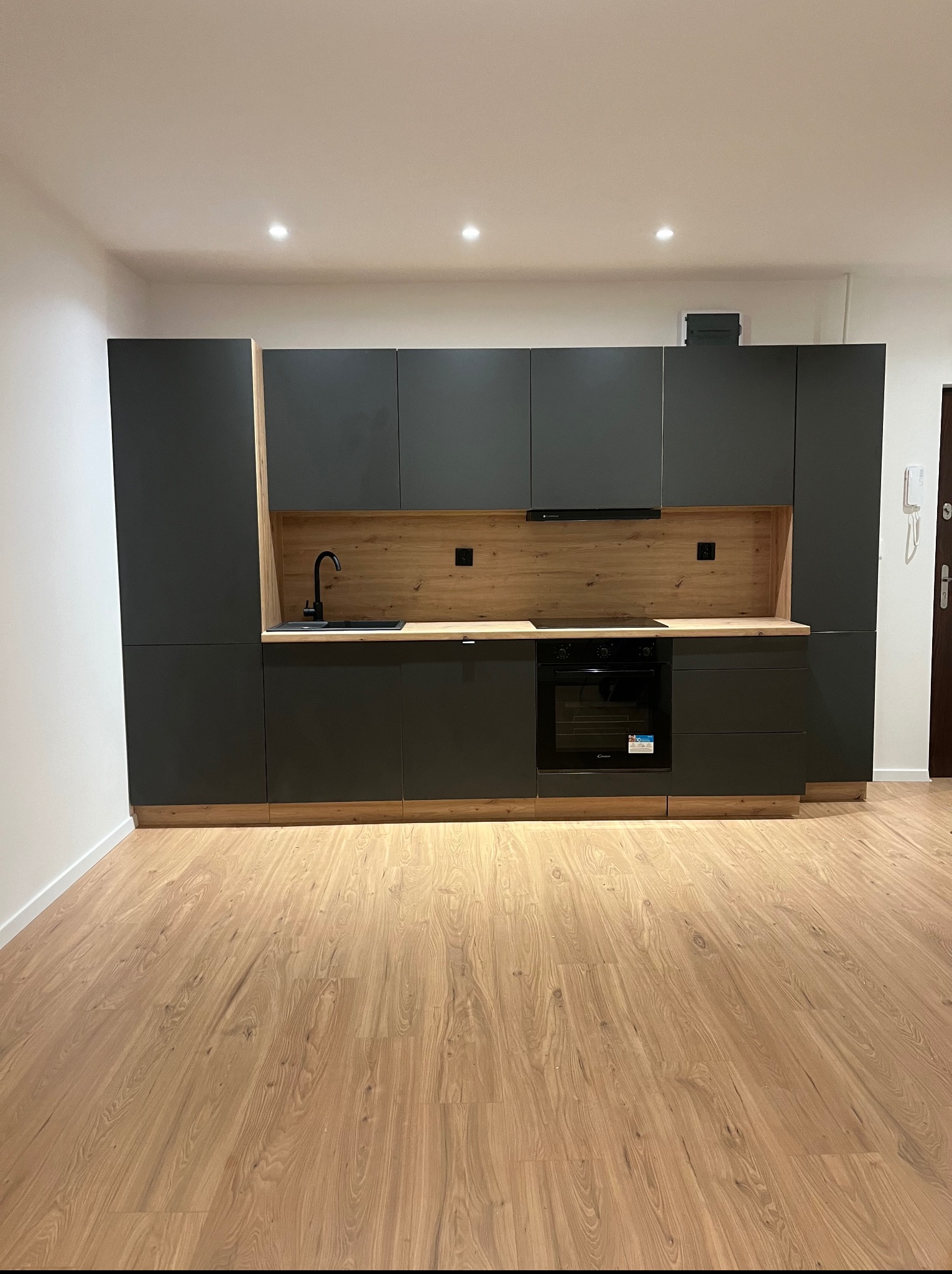 Nowoczesna, szara zabudowa kuchenna z drewnianym akcentem i czarną armaturą, oświetlona punktowo, na tle drewnianej podłogi. Minimalistyczny design.