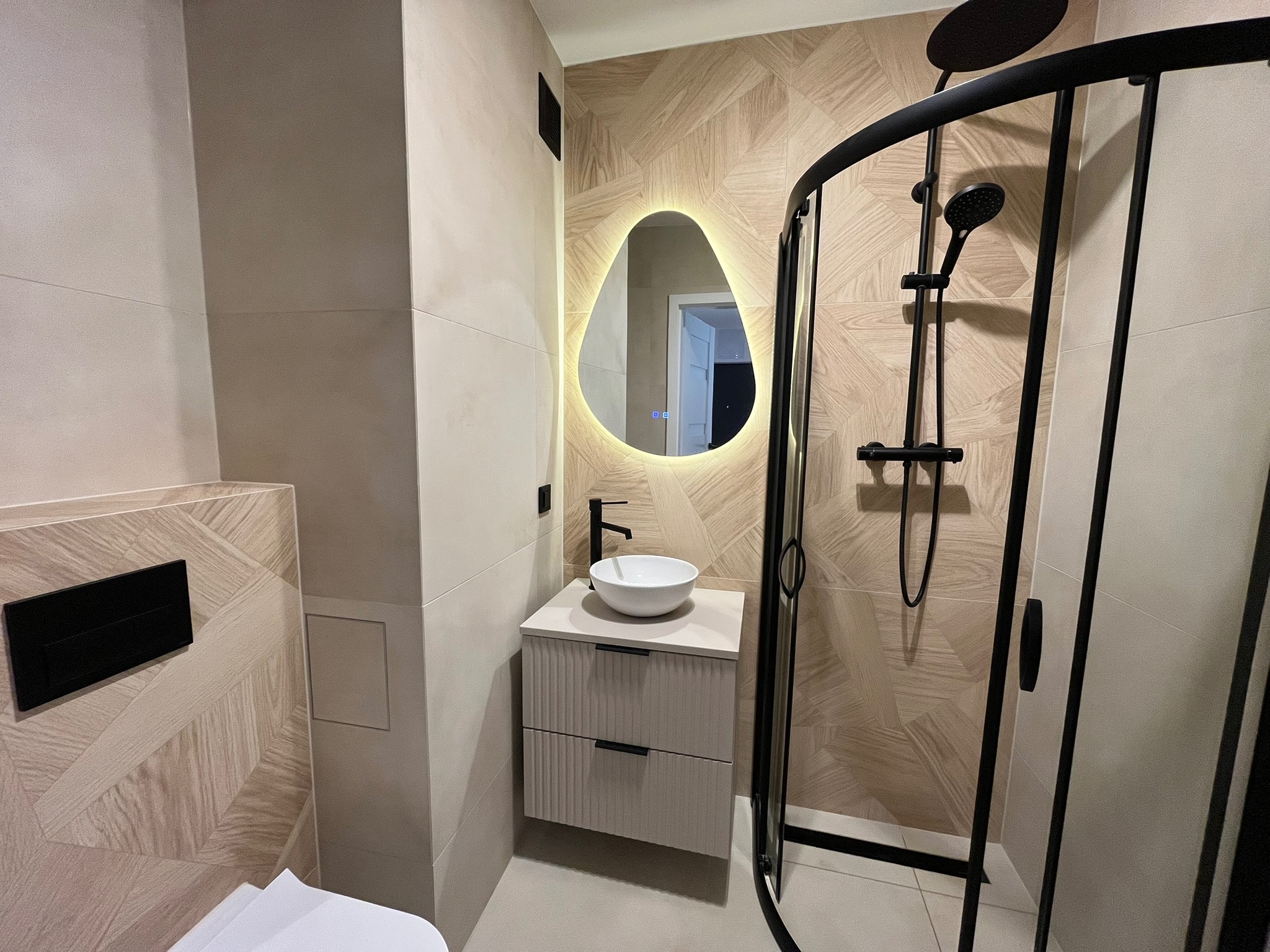Nowoczesna łazienka po remoncie: kabina prysznicowa z czarną armaturą, umywalka na szafce z szufladami i podświetlane lustro o nieregularnym kształcie, beżowe płytki.