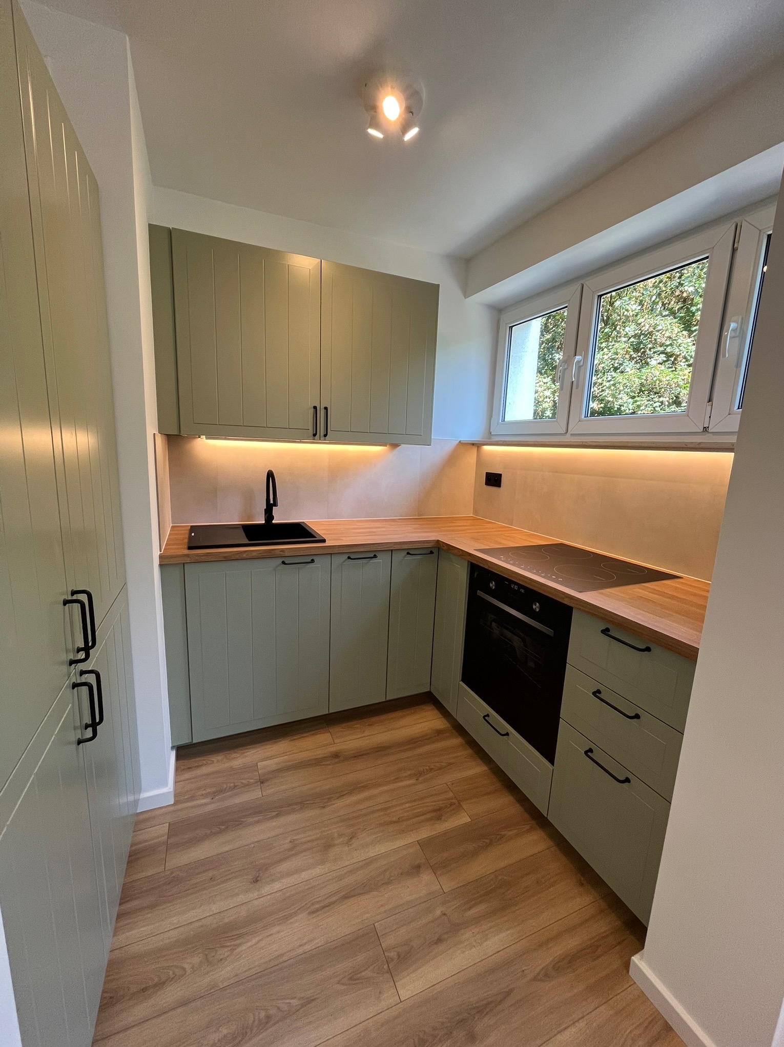 Jasna kuchnia w odcieniach zieleni z drewnianym blatem, czarną armaturą i oświetleniem LED pod szafkami. Widok na okno z zielenią na zewnątrz.