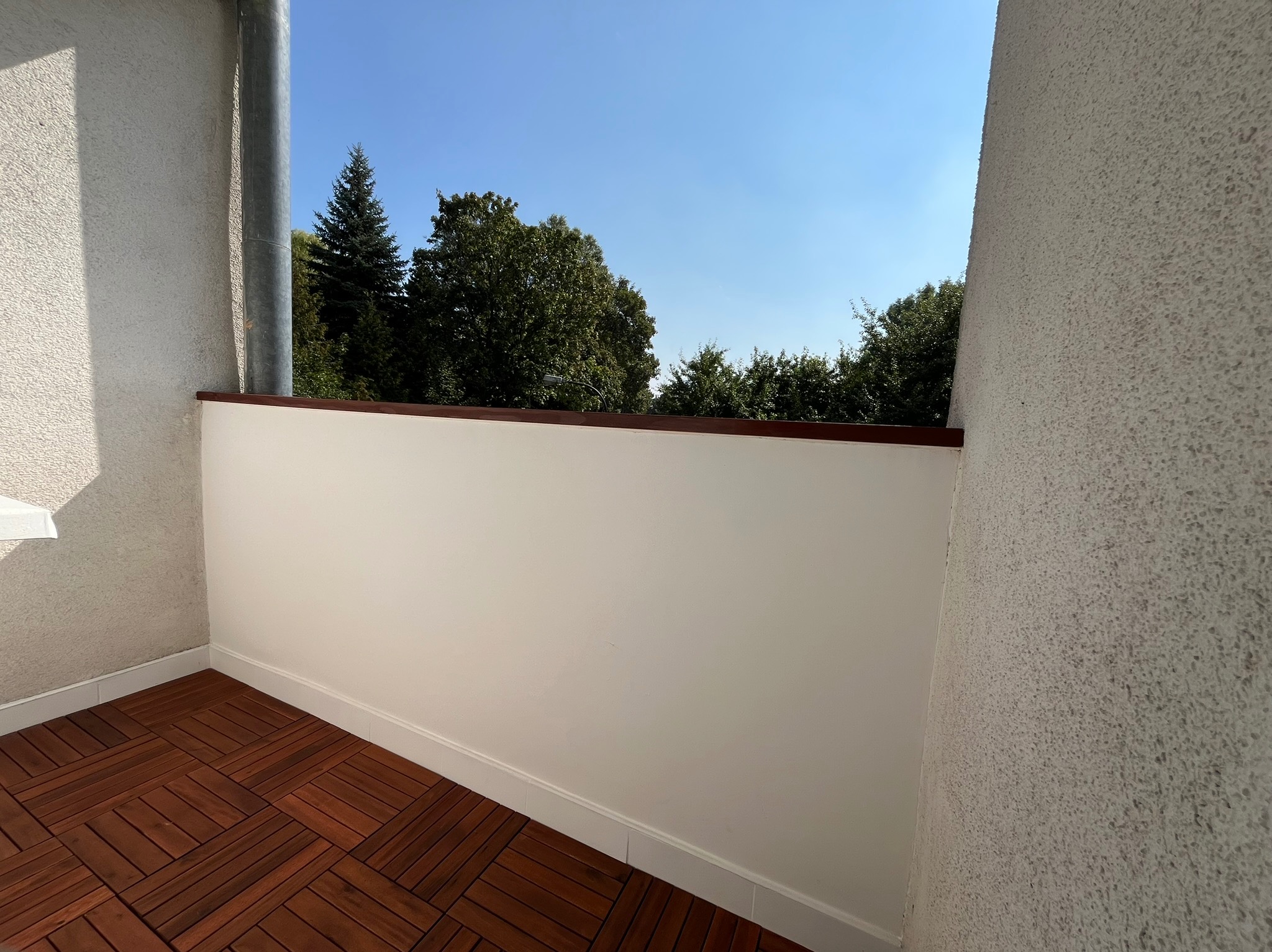 Wykończony balkon z drewnianą podłogą i świeżo pomalowanymi ścianami. Widok na zieleń za balustradą. Słoneczny dzień.