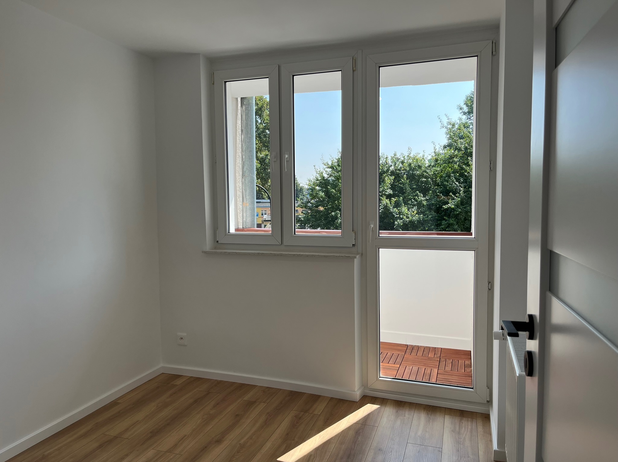 Jasne wnętrze pokoju z drewnianą podłogą i białymi ścianami. Trzyczęściowe okno balkonowe z widokiem na zieleń. Nowoczesne drzwi z czarną klamką po prawej.