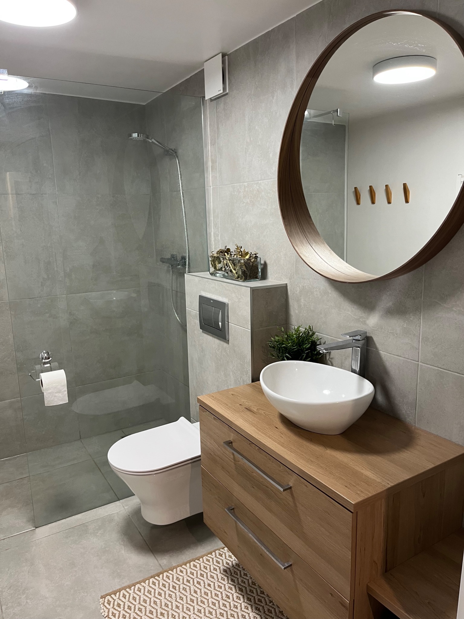 Nowoczesna łazienka z kabiną prysznicową typu walk-in, okrągłym lustrem i drewnianą szafką pod umywalką. Szare płytki i subtelne dekoracje roślinne.