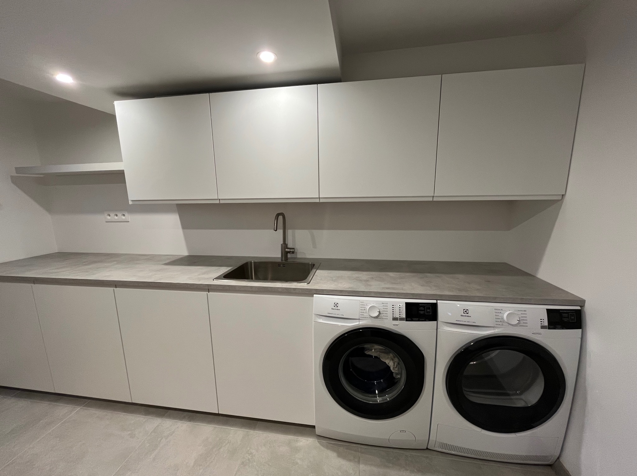 Nowoczesna pralnia w jasnych odcieniach szarości z pralką i suszarką Electrolux w zabudowie. Minimalistyczne szafki wiszące i blat z szarym blatem.