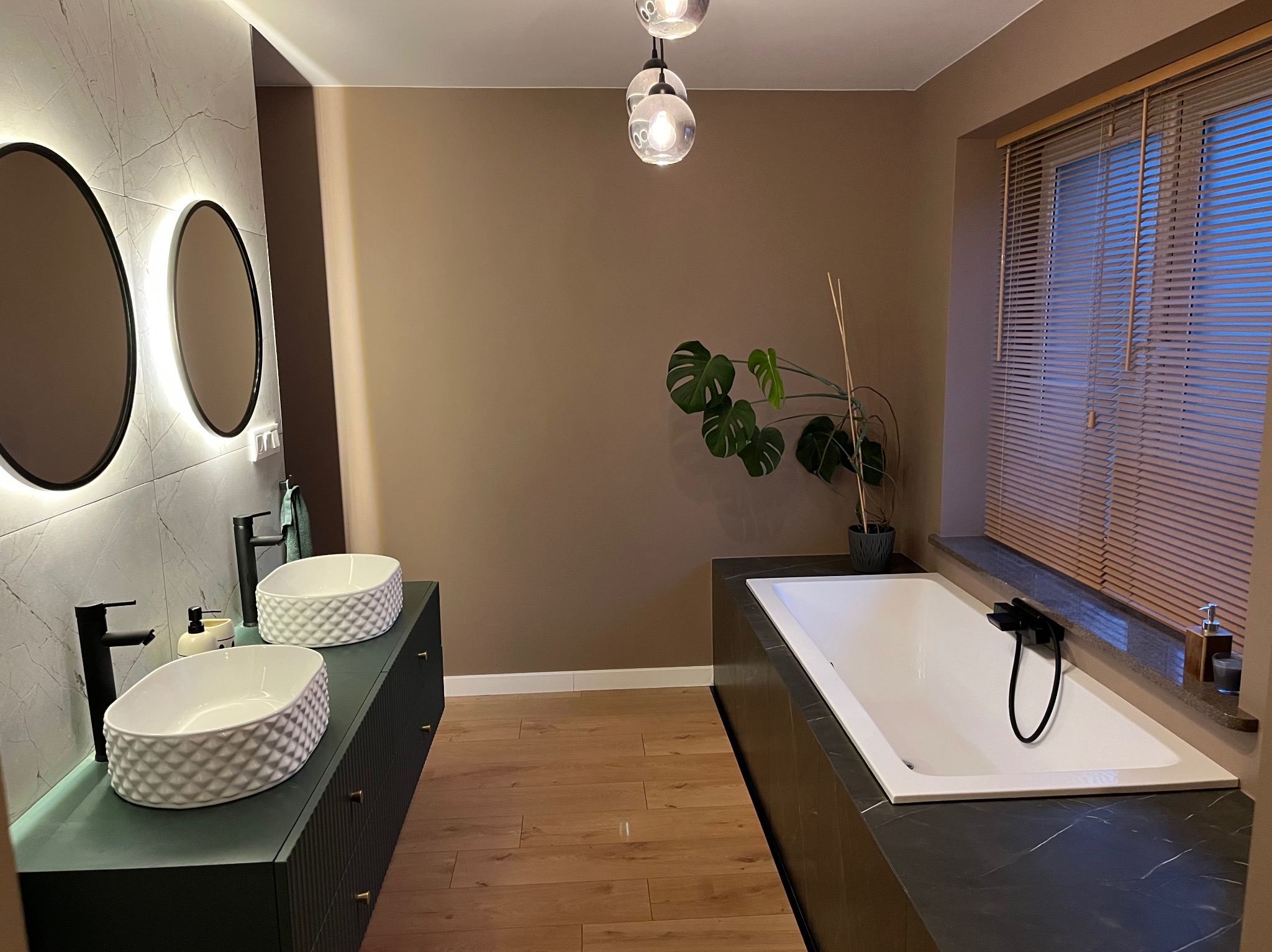 Nowoczesna łazienka z dwoma umywalkami na szafkach w kolorze butelkowej zieleni, owalnymi lustrami z podświetleniem i wolnostojącą wanną.