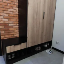 Funkcjonalna szafa z siedziskiem i półkami na buty, w kolorach czerni i drewna, na tle ceglanej ściany. Nowoczesny design.