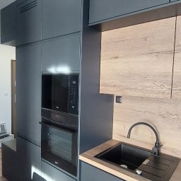 Cenart Filip Cena - Nowoczesna kuchnia z zabudową w odcieniach szarości i drewnianymi akcentami, zintegrowany piekarnik i mikrofalówka, czarny zlew i bateria, drewniana podłoga.