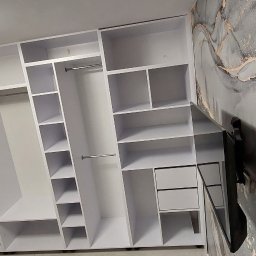 Cenart Filip Cena - Biała szafa wnękowa z półkami i drążkami na ubrania, zamontowana na tle ściany z abstrakcyjnym wzorem. Widoczny fragment telewizora na ścianie.