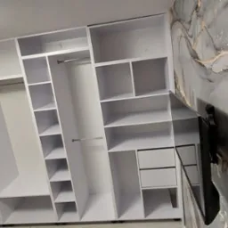 Biała szafa wnękowa z półkami i drążkami na ubrania, zamontowana na tle ściany z abstrakcyjnym wzorem. Widoczny fragment telewizora na ścianie.