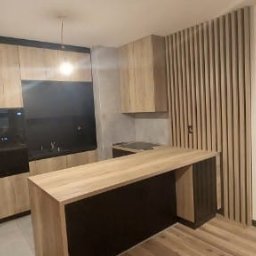 Cenart Filip Cena - Nowoczesna kuchnia z drewnianymi frontami i czarnymi akcentami, wyspa kuchenna, pionowe lamele na ścianie, widok na toaletę w tle.