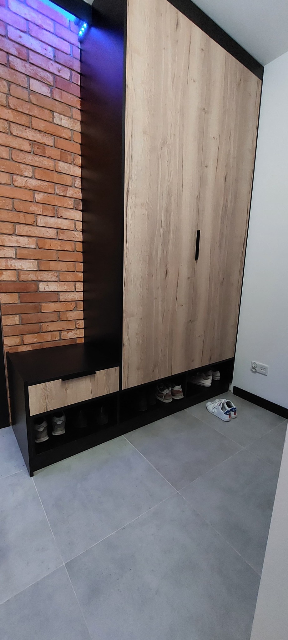Funkcjonalna szafa z siedziskiem i półkami na buty, w kolorach czerni i drewna, na tle ceglanej ściany. Nowoczesny design.