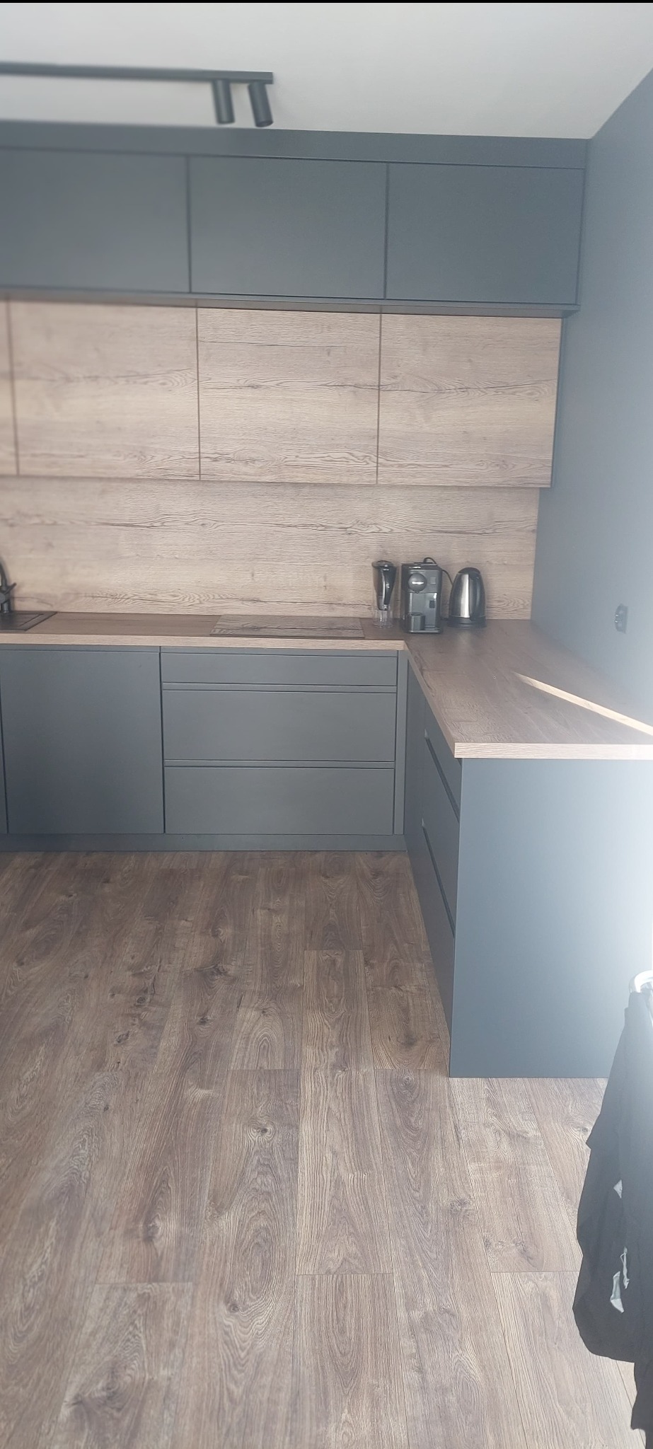 Nowoczesna kuchnia z szarymi szafkami i drewnopodobnym blatem, oświetlona punktowo. Widoczna płyta indukcyjna i ekspres do kawy. Podłoga z paneli.