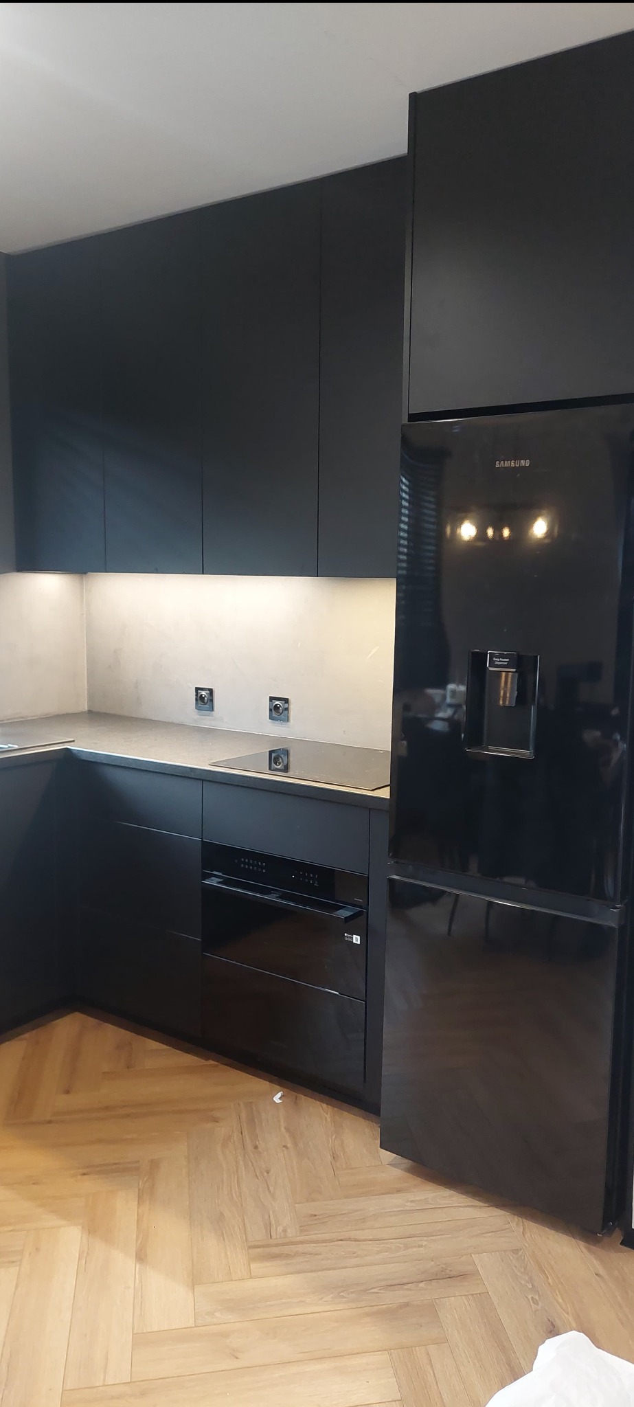 Nowoczesna, czarna kuchnia na wymiar z zabudowaną lodówką Samsung i drewnianą podłogą ułożoną w jodełkę, oświetlenie podszafkowe.