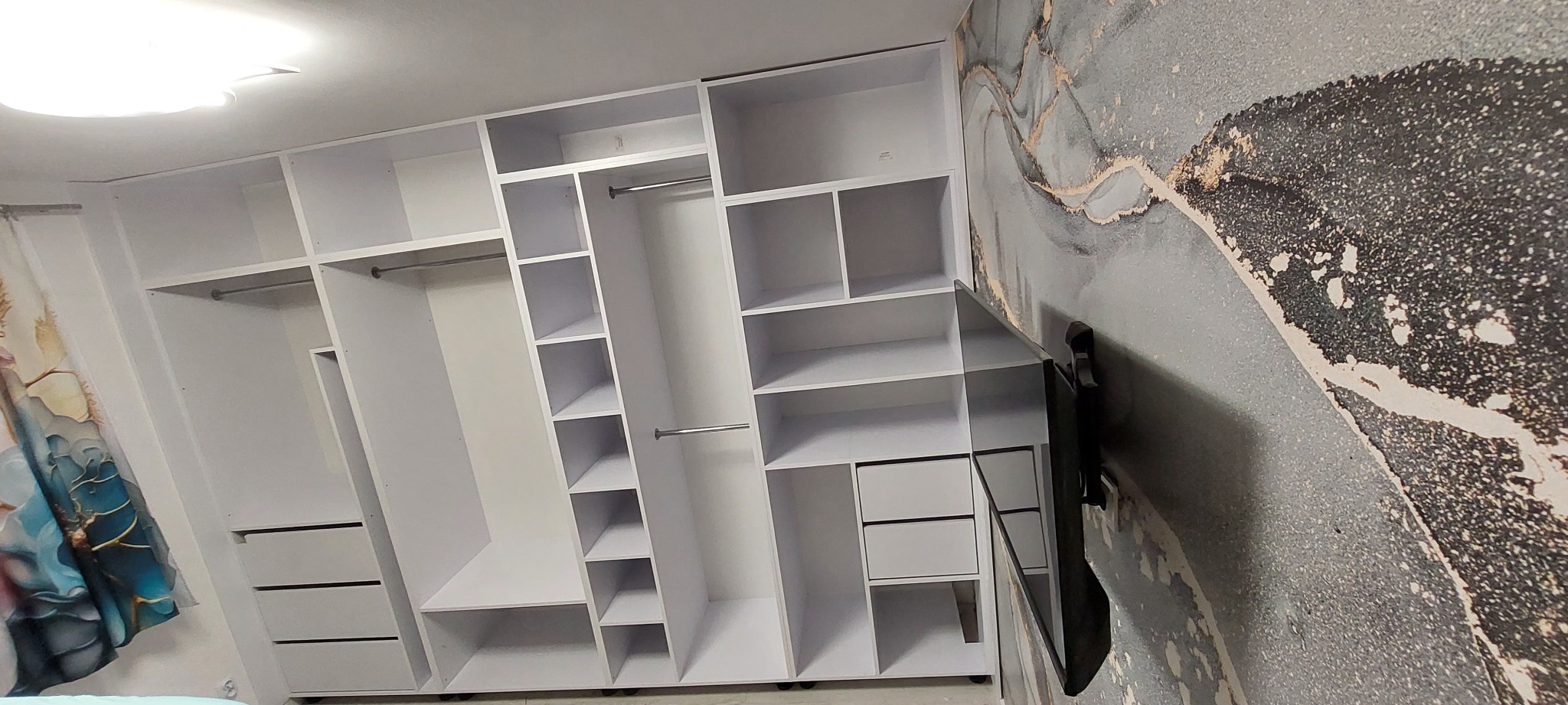 Biała szafa wnękowa z półkami i drążkami na ubrania, zamontowana na tle ściany z abstrakcyjnym wzorem. Widoczny fragment telewizora na ścianie.