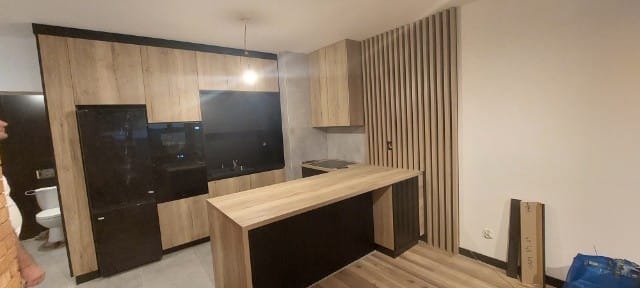 Nowoczesna kuchnia z drewnianymi frontami i czarnymi akcentami, wyspa kuchenna, pionowe lamele na ścianie, widok na toaletę w tle.