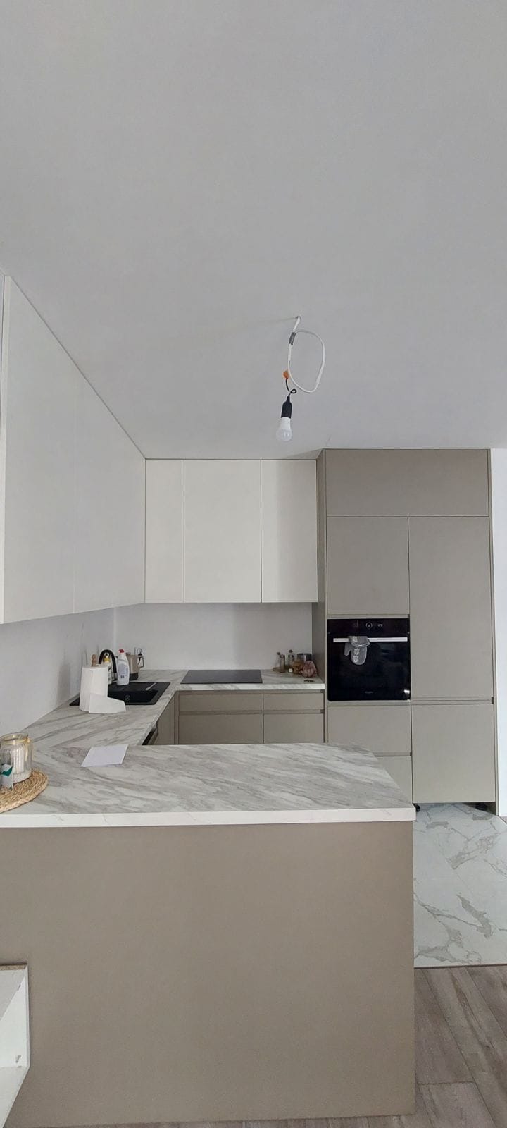 Jasna, nowoczesna kuchnia z marmurowym blatem i szarymi szafkami. Widoczny piekarnik w zabudowie i minimalistyczny design. Uwagę przyciąga surowa żarówka na kablu.