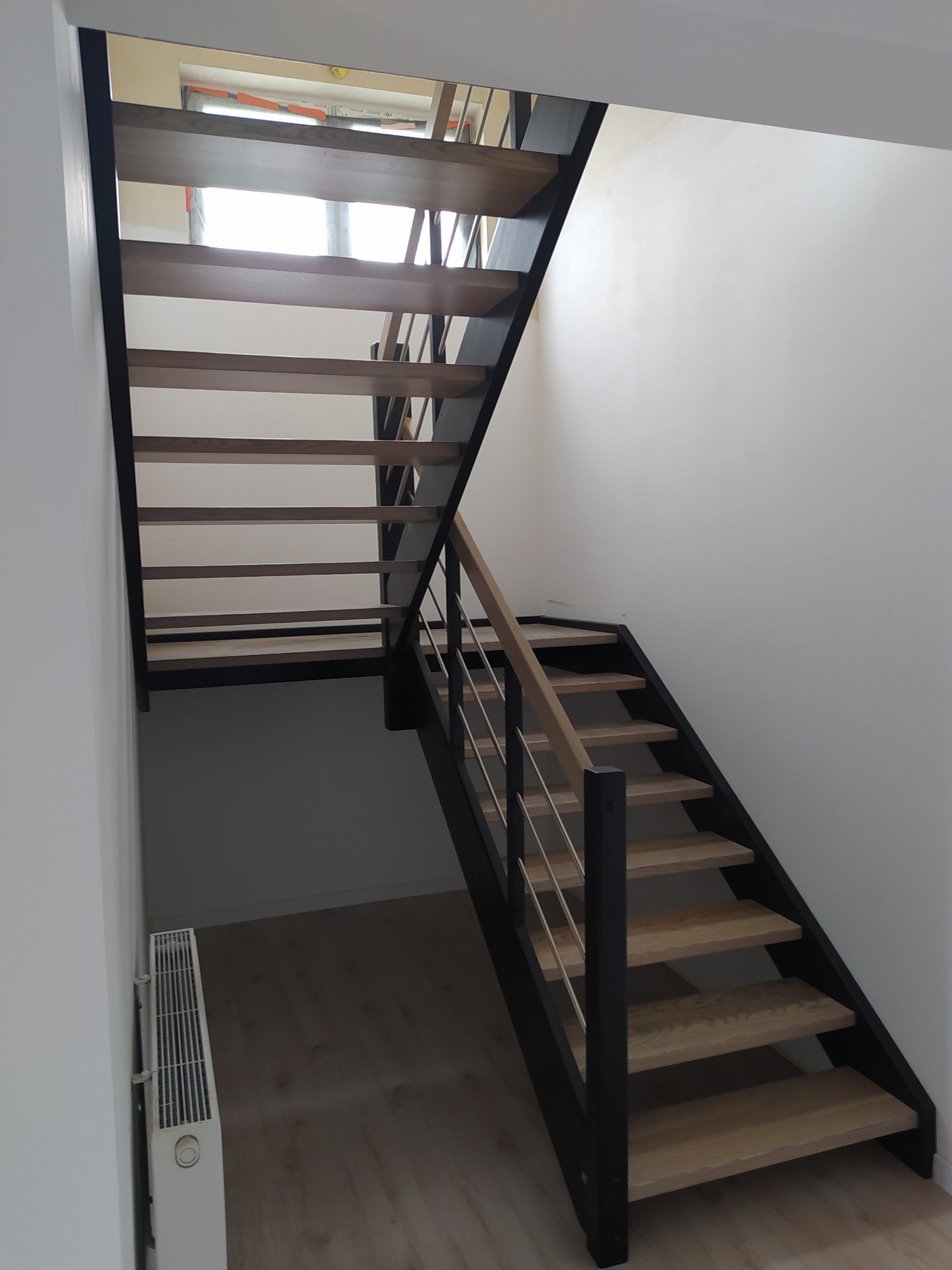 Nowoczesne schody z drewnianymi stopniami i czarną metalową konstrukcją, z jasną drewnianą balustradą i metalowymi prętami, w jasnym wnętrzu.