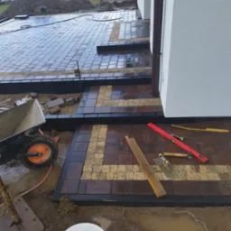 Układanie kostki brukowej w dwóch kolorach, z widocznymi narzędziami pracy i fragmentem elewacji budynku. W tle ziemia i zieleń, etap prac w toku.