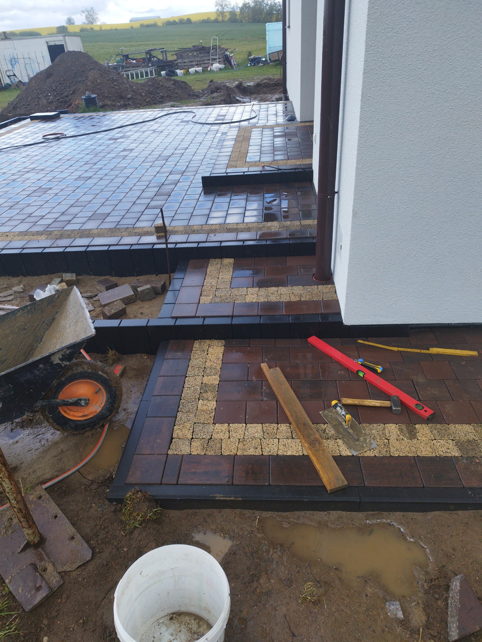 Układanie kostki brukowej w dwóch kolorach, z widocznymi narzędziami pracy i fragmentem elewacji budynku. W tle ziemia i zieleń, etap prac w toku.