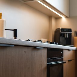 Wojciech Psiuk Manufaktura Mebli - Nowoczesna kuchnia: zabudowa z drewnianymi frontami, czarny piekarnik i oświetlenie LED pod szafkami. Minimalistyczny design z ekspres do kawy w tle.