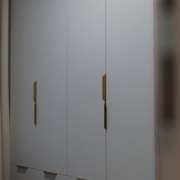 Wojciech Psiuk Manufaktura Mebli - Elegancka, szara szafa wnękowa z złotymi, minimalistycznymi uchwytami, podzielona na część z drzwiami i dolne szuflady. Widoczne drewniane wykończenie boku szafy.