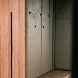 Wojciech Psiuk Manufaktura Mebli - Drewniana szafa z długim, czarnym uchwytem sąsiaduje z tapicerowaną wnęką z siedziskiem i wieszakami. Nowoczesny design, stonowana kolorystyka.