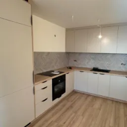 Jasna kuchnia z frontami w odcieniach beżu i szarości, drewniany blat i podłoga, wzorzysta ściana nad blatem. Widoczne płyty grzewcze i piekarnik.