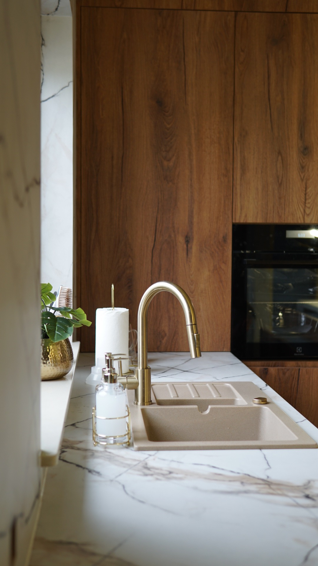 Beżowy zlew z baterią w kolorze złota na blacie z marmurowym wzorem. W tle drewniane fronty szafek kuchennych i piekarnik. Nowoczesna kuchnia.